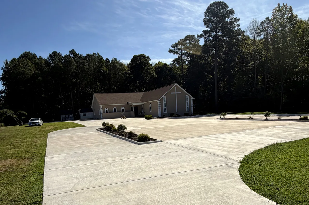 A wide paved drive and parking lot in daylight, with landscaped islands and lawn on both sides, leading to the venue building among tall trees