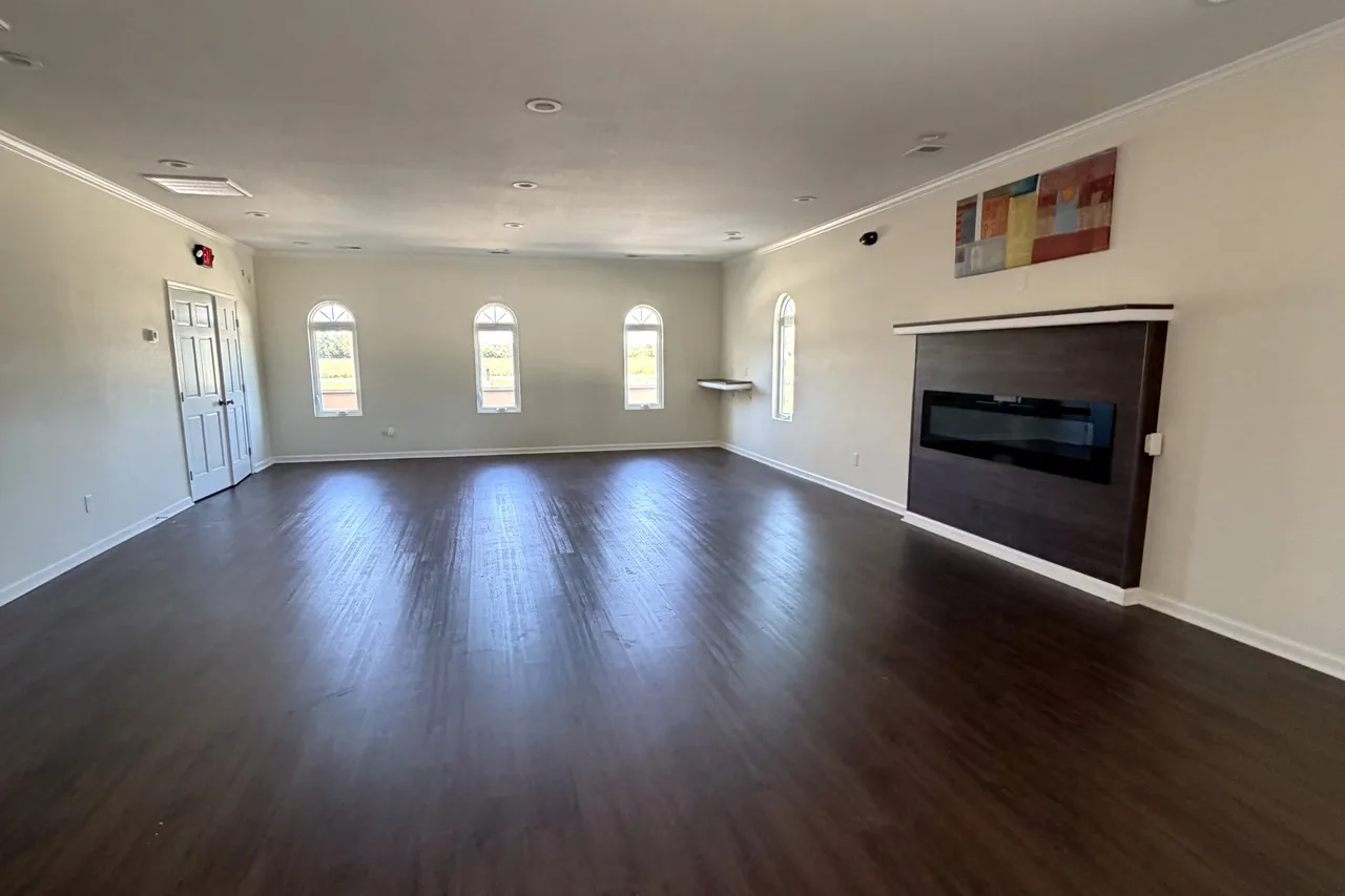 The Fireside Room: an open room with dark wood-look floors, arched windows along the far wall, a dark fireplace feature wall on the right, double doors on the left, and recessed ceiling lights