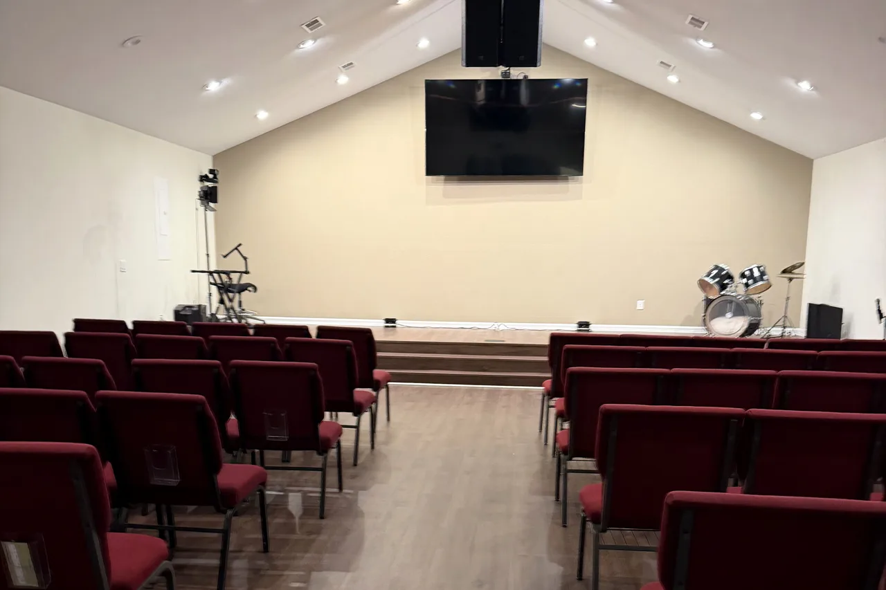 The raised stage at the front of The Stage Hall, with steps up from the floor, a large wall-mounted screen, and rows of red upholstered chairs in front