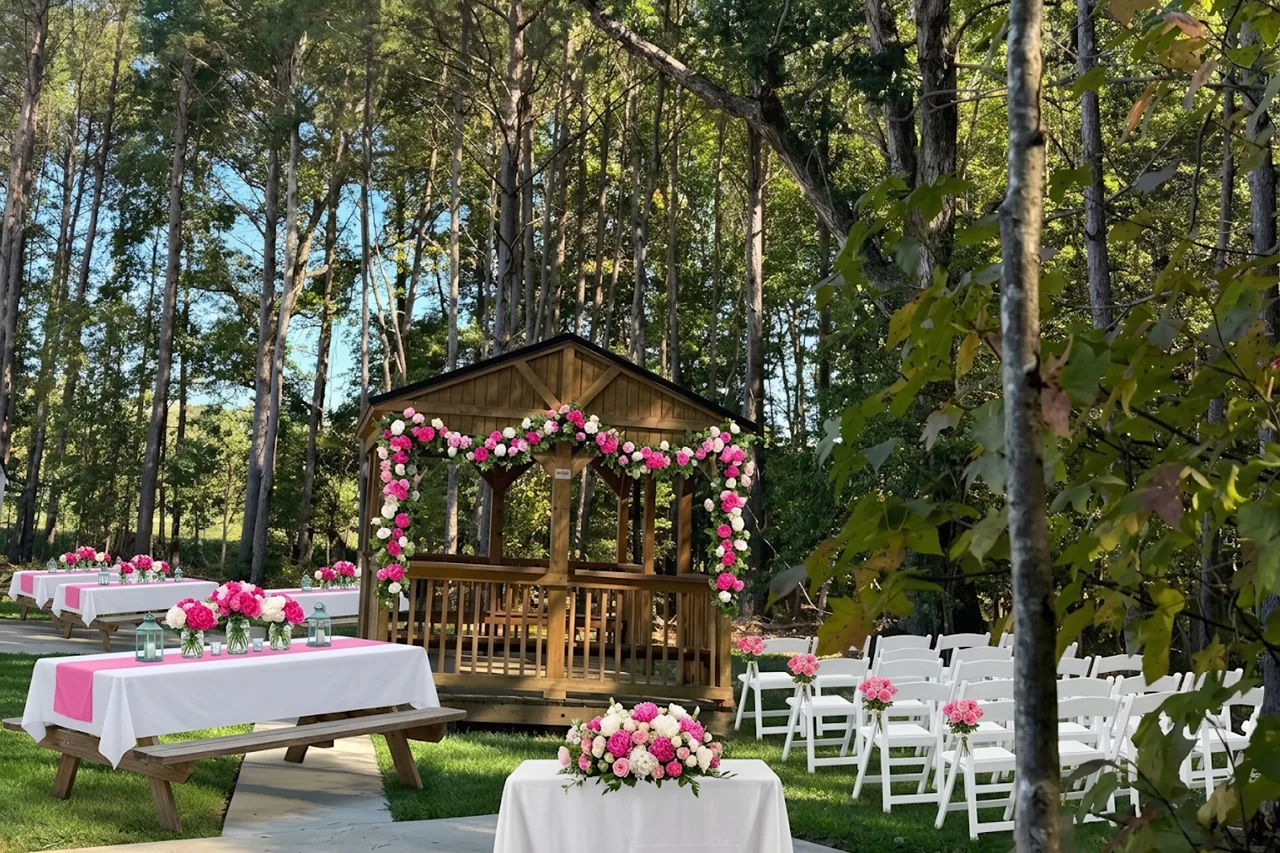 Styled concept: a community gathering in The Pine Garden, with picnic tables in white and pink, rows of white chairs on the lawn, a welcome table with pink and white roses, and rose garlands on the gazebo